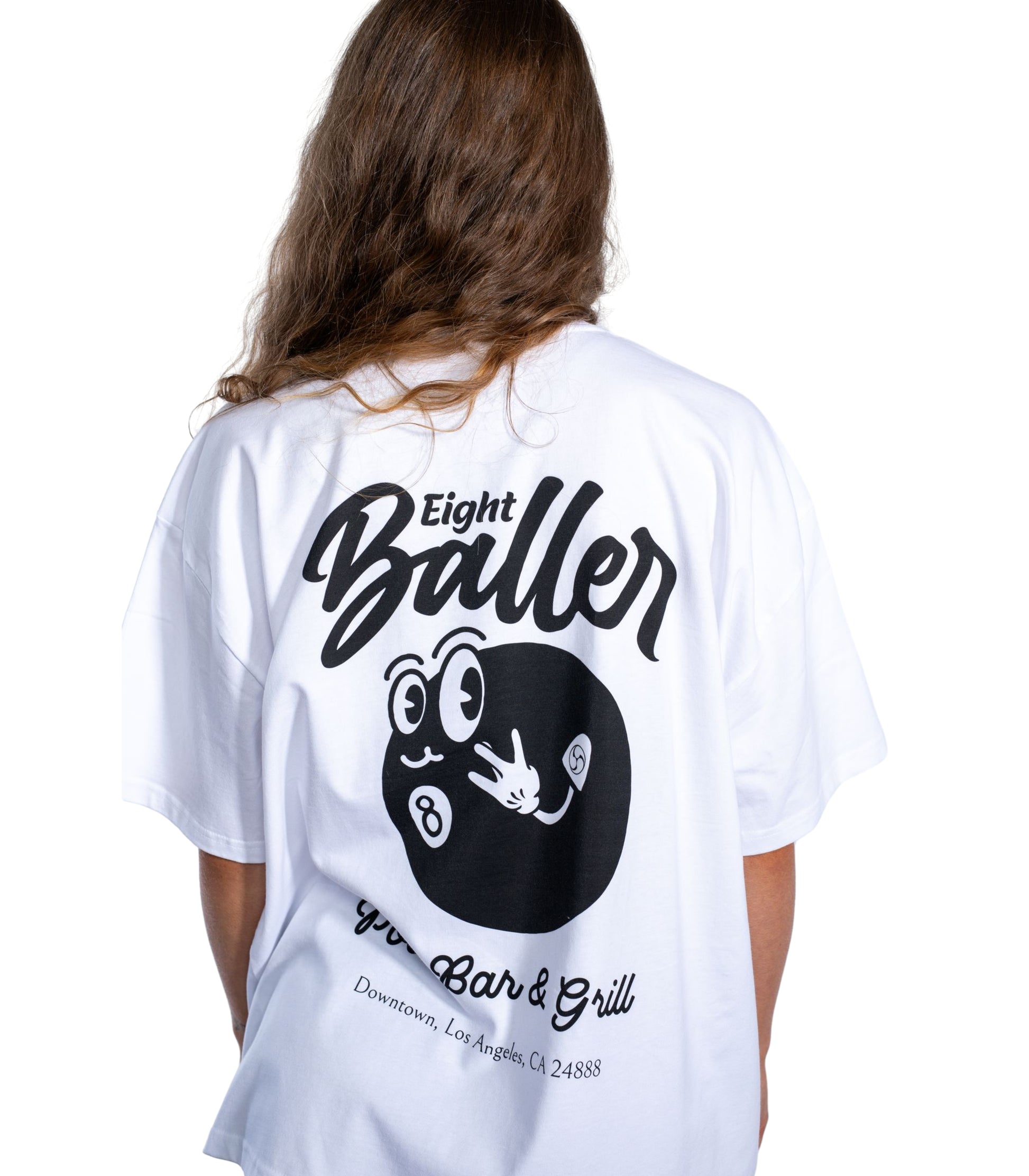 Back view of woman wearing oversized white Flow Like Zen streetwear t-shirt featuring large Eight Baller cartoon 8-ball back print and 'Pool Bar & Grill, Downtown Los Angeles' slogan – UK independent fashion.