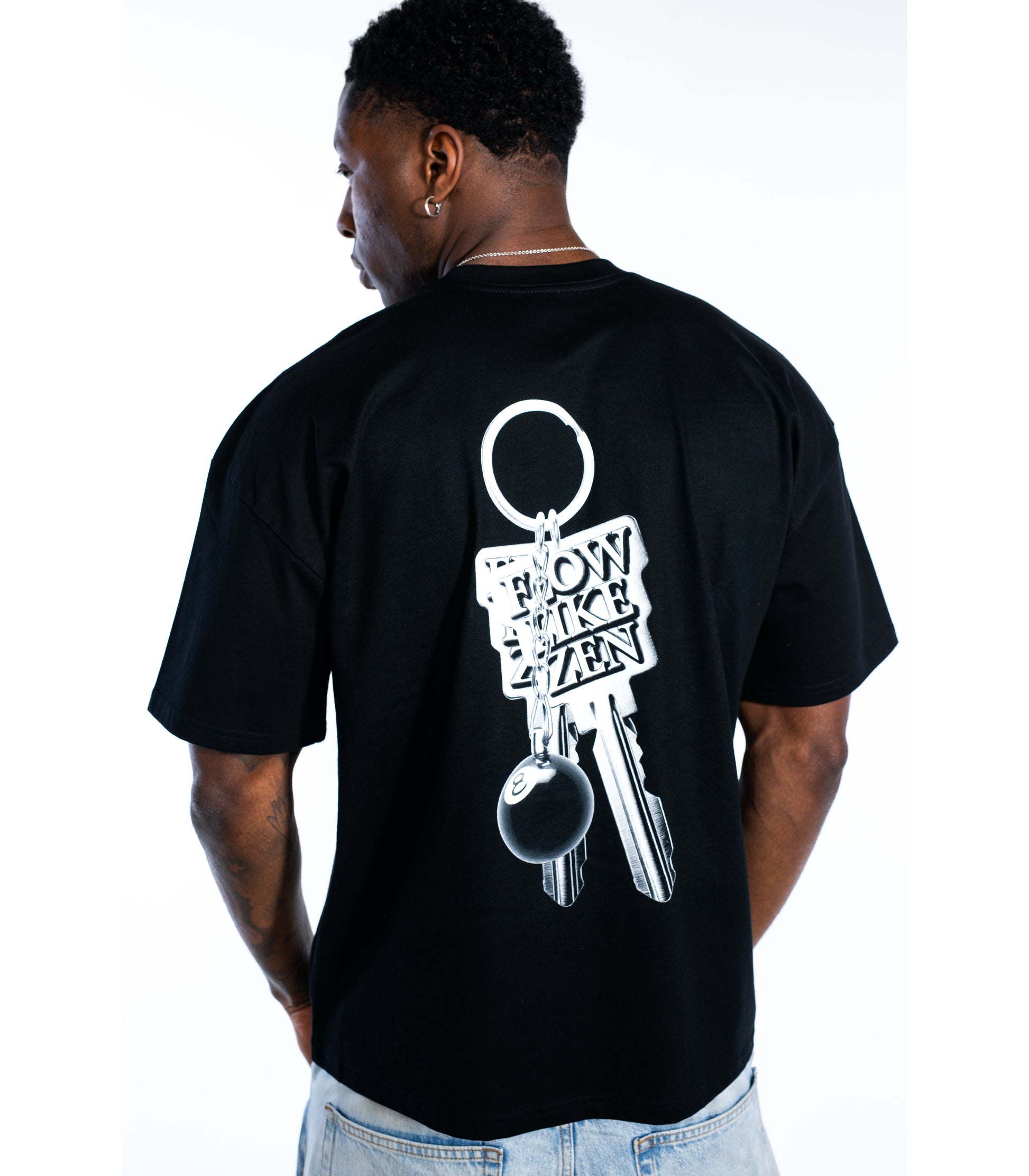 Back view of man wearing black Flow Like Zen streetwear t-shirt featuring metallic keychain back graphic with 8-ball and 'Flow Like Zen' logo – UK skatewear and urban fashion.