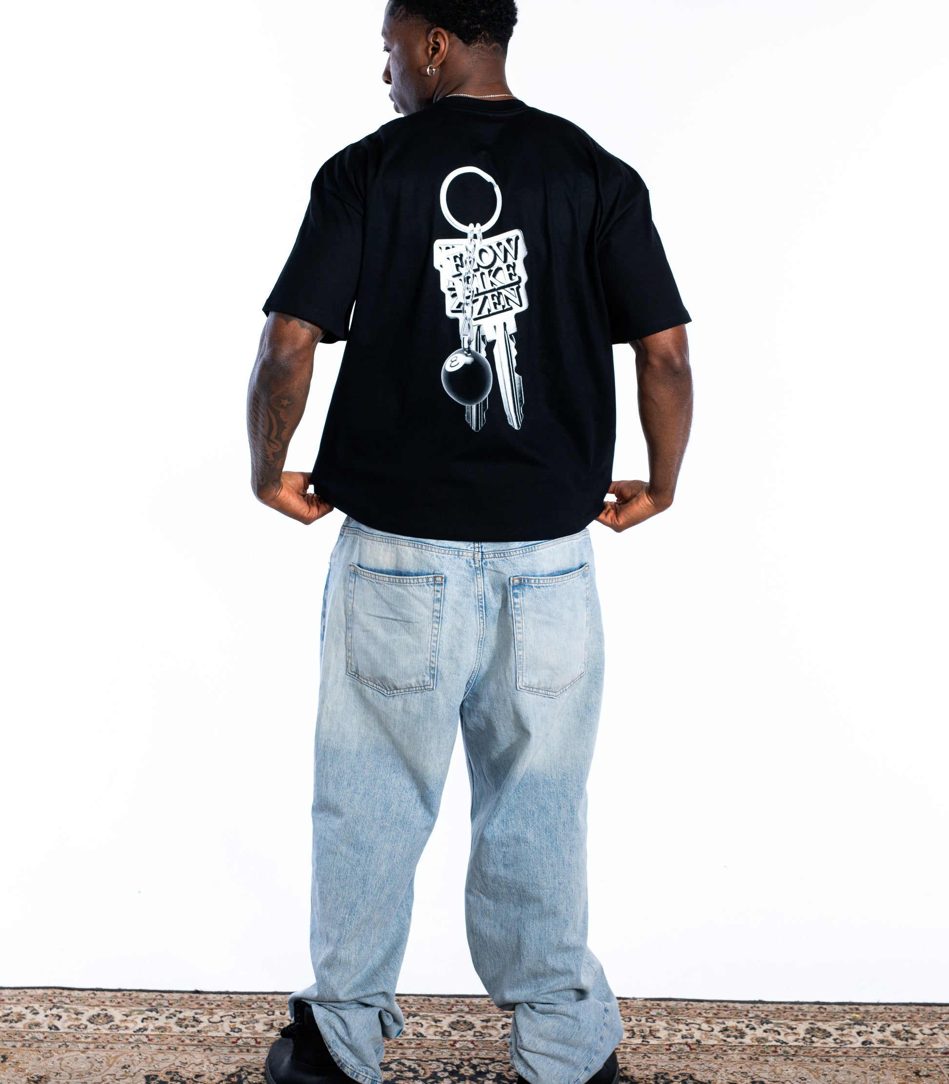Back view of male model wearing black Flow Like Zen t-shirt with large silver keychain back graphic and 8-ball – UK skatewear and urban fashion brand.