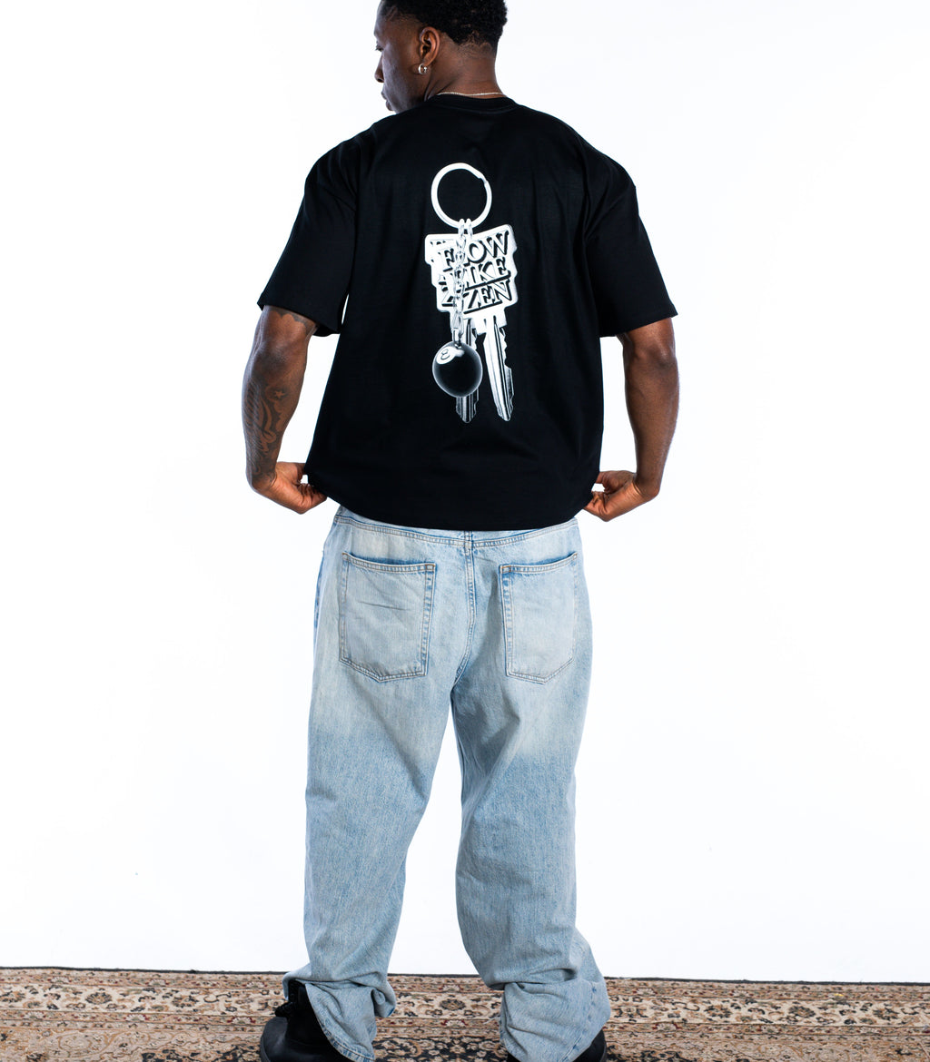 Back view of male model wearing black Flow Like Zen t-shirt with large silver keychain back graphic and 8-ball – UK skatewear and urban fashion brand.